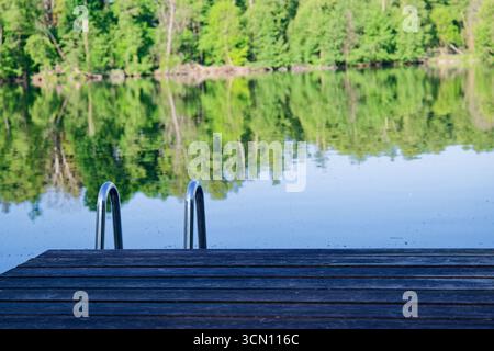 Un semplice molo in legno si estende sopra l'acqua ferma del lago. Due guide metalliche da una scala conducono nelle basse profondità. I riflessi degli alberi ondulano dolcemente, e il Foto Stock