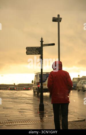 LONDRA, REGNO UNITO. 19 OTTOBRE 2010. Una figura in un impermeabile rosso si erge su un ponte di Londra bagnato dalla pioggia al tramonto, stagliato contro il suggestivo cielo dorato Foto Stock