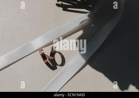 Due fedi nuziali dorate poggiano su una superficie bianca, circondate da un nastro di raso e da ombre spettacolari, catturando l'essenza dei momenti di matrimonio e dell'unità. Foto Stock