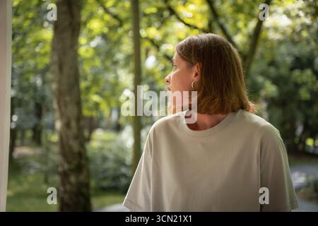 Una giovane donna con una t-shirt grigia chiaro guarda attentamente in lontananza mentre si trova in una foresta dondolata dal sole Foto Stock