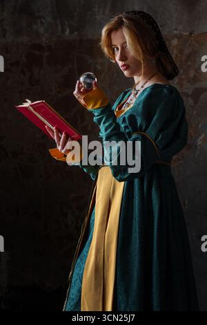 Una bella signora nobile in un abito rinascimentale color smeraldo con un libro e una sfera di cristallo nelle sue mani. Una giovane bionda in costume medievale, ritratto sullo sfondo di un muro di pietra del castello Foto Stock