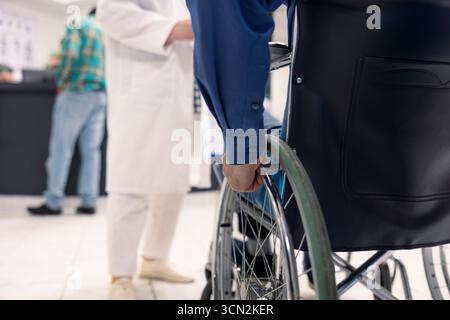 Mano di persona bianca che afferrano la sedia a rotelle nella hall dell'ospedale. Paziente di sesso maschile caucasico con disabilità che si prepara per il controllo medico con operatori sanitari in sala d'attesa clinica. Foto Stock