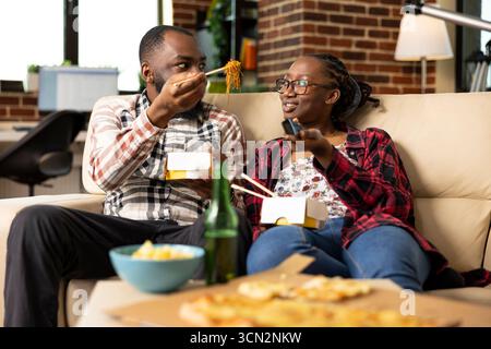 Coppia afroamericana che si rilassa a casa, condividendo spaghetti da asporto durante la serata romantica. L'uomo nutre la sua ragazza con le bacchette mentre si gode del tempo di qualità sul comodo divano dell'appartamento. Foto Stock