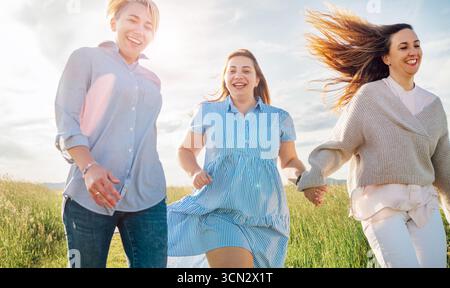 Ritratto a basso angolo di quattro donne allegre che si tengono per mano e corrono su un prato sotto il cielo blu con nuvole, simboleggiando amicizia, gioia, unità, un Foto Stock