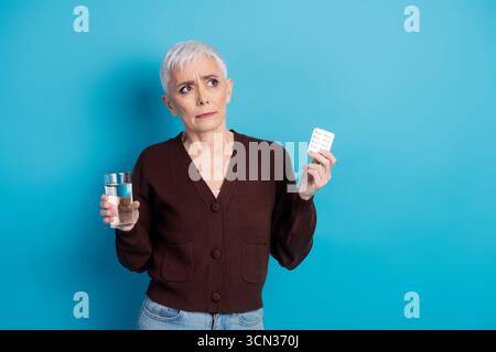 Donna anziana che tiene in mano acqua e farmaci mentre mostra espressioni riflessive su sfondo blu Foto Stock