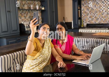 Madre e figlia indiane che scattano selfie con lo smartphone mentre utilizzano il computer portatile al tavolo della cucina Foto Stock