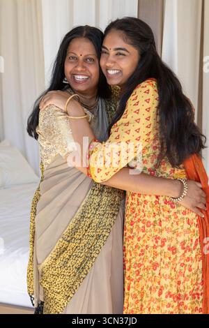 Madre e figlia indiane in abiti tradizionali abbracciati, sorridenti in camera da letto con gioielli, bindi Foto Stock