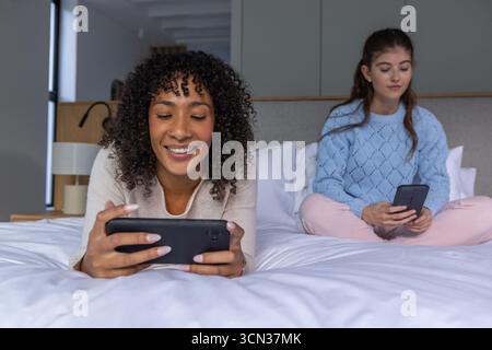 Diverse amiche che si rilassano sul letto bianco in camera da letto, utilizzano smartphone, spazio di copia Foto Stock