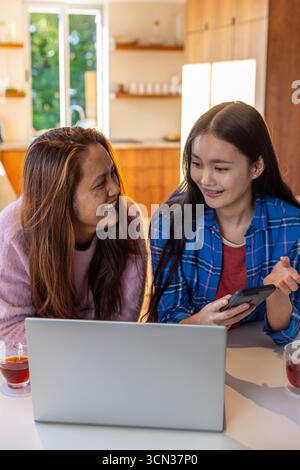 Madre e figlia asiatiche che usano smartphone e laptop argento al tavolo della cucina con tè Foto Stock