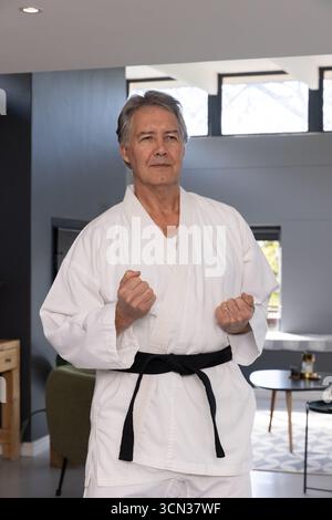 Uomo maturo in piedi nel soggiorno con indosso la cintura bianca e nera che tiene i pugni fissati Foto Stock