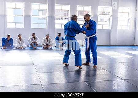 Istruttore afroamericano che afferra lo studente gi, dimostrando jiu-jitsu su tappetini dojo Foto Stock