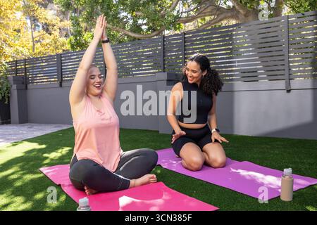 Diverse amiche di abbigliamento sportivo che praticano yoga in un cortile illuminato dal sole sui tappetini Foto Stock