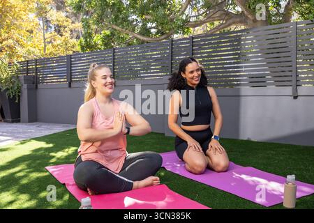 Diverse amiche che praticano yoga sul tappeto erboso tramite recinzione a lastre utilizzando tappetini e bottiglie d'acqua Foto Stock