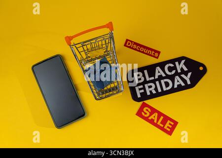 Carrello della spesa in miniatura con confezione regalo e smartphone su sfondo giallo con etichette Black friday e sconti per promuovere lo shopping e le vendite online Foto Stock