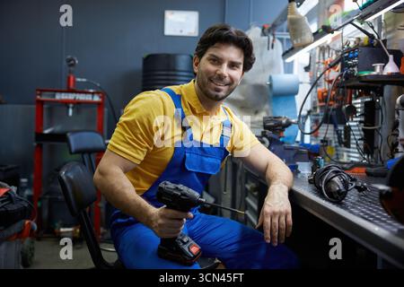 Un lavoratore siede comodamente su una sedia, tenendo un trapano e sorridendo Foto Stock
