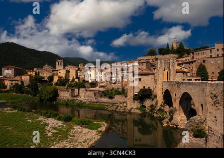 A Besalú, un'antica città catalana di Girona, in Spagna, il fiume Fluvià costeggia i resti di mura difensive dopo essere passato sotto gli archi arrotondati del Viejo o Ponte Vecchio dell'XI secolo ricostruito. La passerella del ponte, lunga 105 m o 344 piedi, termina in una porta all'estremità della città Besalú è una delle città medievali meglio conservate della Catalogna, con molte chiese storiche e altri edifici. Foto Stock