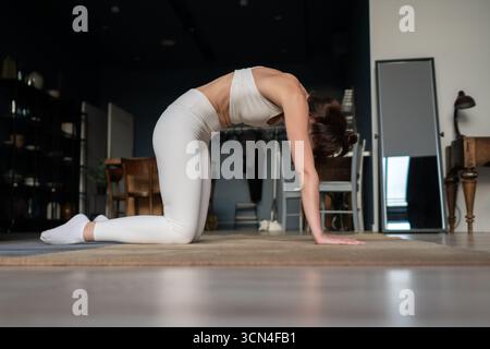 Una donna in abbigliamento sportivo bianco esegue una posa di gatto yoga in interni, con l'arco della schiena verso l'alto su mani e ginocchia, pratica la mobilità spinale, il coinvolgimento del nucleo, h Foto Stock