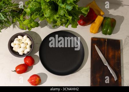 Un ripiano pulito della cucina mostra un piatto nero circondato da pomodori freschi, peperoni colorati, lattuga e mozzarella. Un coltello poggia su un asse di legno, pronto per la preparazione dei pasti. Foto Stock