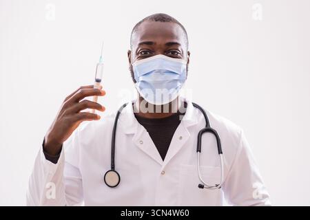 Ritratto del medico afroamericano con maschera protettiva che tiene la siringa e guarda la fotocamera su sfondo grigio. Foto Stock