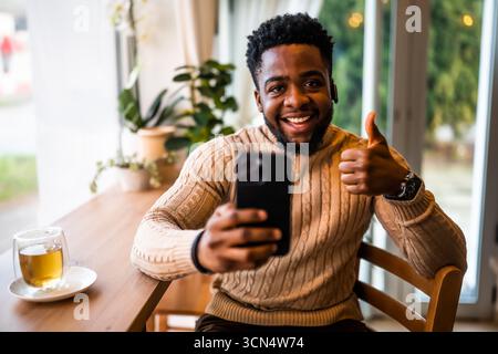 Un uomo nero sorridente siede al bar, vestito in modo informale. Sta utilizzando smartphone e messaggistica online. Foto Stock