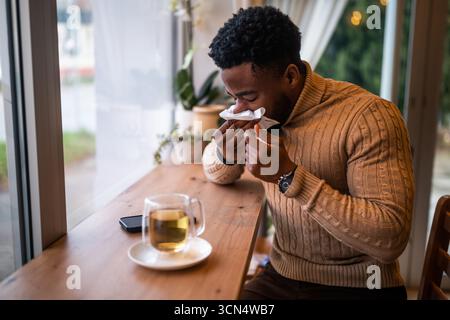 Giovane seduto al bar, rilassandosi e bevendo tè. Sta soffiando il naso. Foto Stock