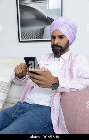 Uomo indiano medio adulto che indossa lo smartphone turban mentre è seduto in soggiorno Foto Stock