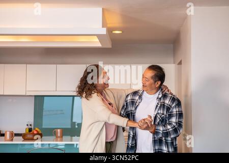 Diverse coppie anziane che ballano tenendosi per mano in cucina vicino al piano di lavoro con un cestino di frutta Foto Stock