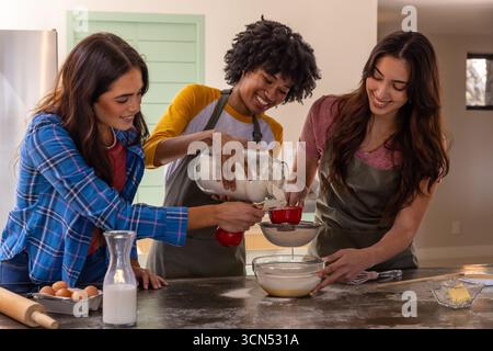 Diverse amiche che misurano la farina con una tazza e frullano nella ciotola di Kitchen Island Foto Stock