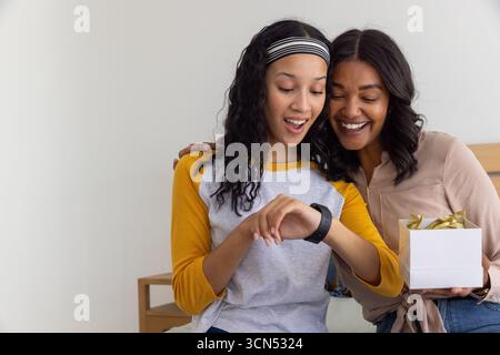 Mamma e figlia adolescente diverse sedute sulla panchina in camera da letto a esaminare smartwatch e confezione regalo Foto Stock
