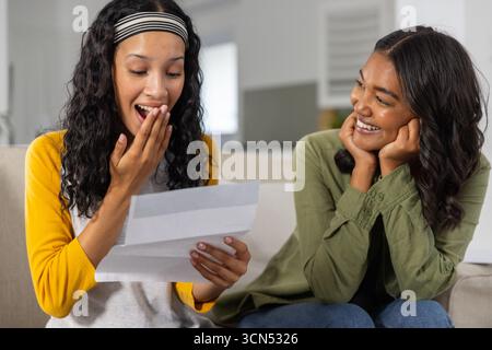 Mamma e figlia adolescente diverse sedute sul divano in soggiorno a leggere una lettera sorridente Foto Stock