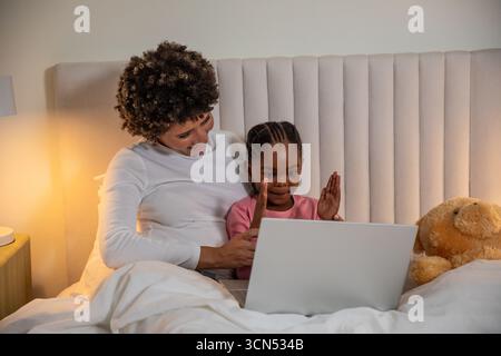 Madre ispanica con ragazza afro-americana che condivide un laptop argentato con biancheria bianca Foto Stock