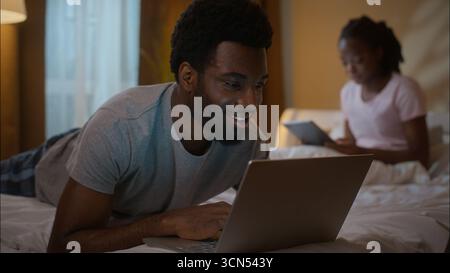 Famiglia afroamericana marito che utilizza laptop moglie tablet digitale camera da letto sera coppia etnica rilassarsi sdraiato letto fine settimana gadget dipendenza scorrimento Foto Stock
