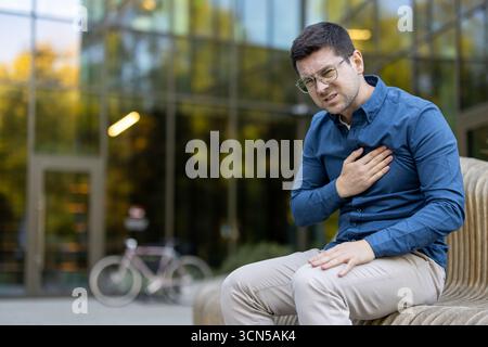 Giovane che soffre di dolore toracico acuto e disagio, seduto su una panchina all'esterno di un edificio moderno, mostrando sintomi di un potenziale attacco di cuore o emergenza cardiaca Foto Stock