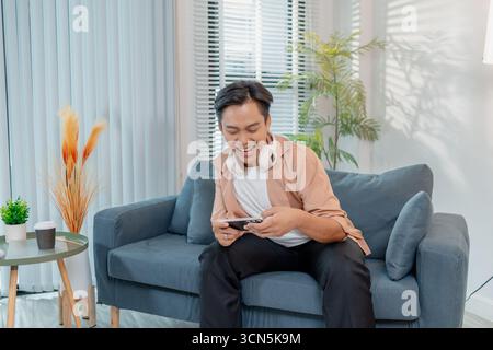 Un giovane uomo si siede su un divano a casa con le cuffie intorno al collo, tenendo in mano uno smartphone e giocando a un gioco mobile, sorridendo e festeggiando con l'accisa Foto Stock