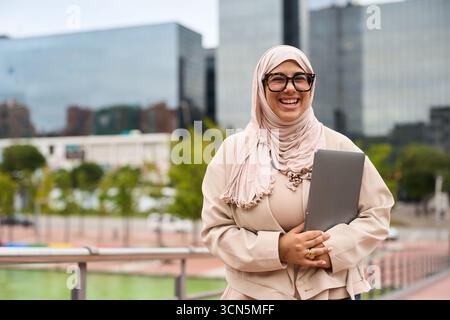 Donna d'affari musulmana che indossa hijab e occhiali sorridenti in città Foto Stock