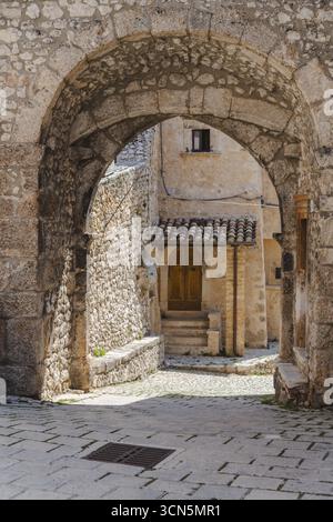 Vista dell'arco in pietra intemprata che incornicia un cortile soleggiato con una porta in legno e gradini, creando una scena senza tempo, Santo Stefano di Sessanio, A. Foto Stock