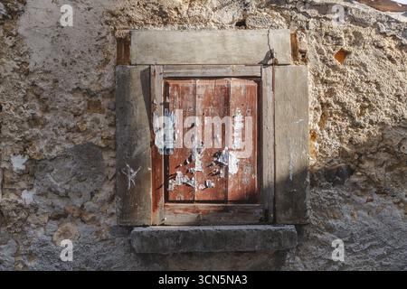 Veduta di una finestra a bordo, rossa tenue, segnata da cartelli sbucciati e incorniciata da pietre sbriciolate e strutture in cemento, Calascio, Abruzzo, Italia. Foto Stock