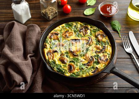 Frittata con spinaci e funghi champignon in padella di ghisa su un tavolo di legno marrone. Vista ravvicinata Foto Stock