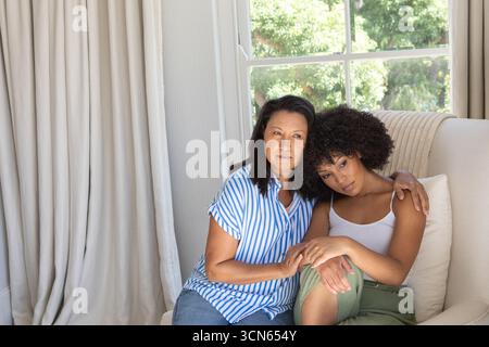 Madre asiatica e figlia afroamericana seduta sul divano con cuscino a casa e tenuta per mano Foto Stock