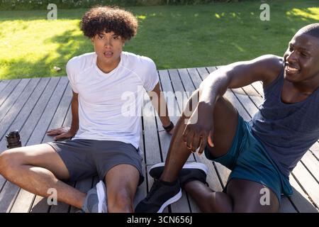 Amici maschi diversi che riposano sul ponte dopo l'allenamento con bottiglia d'acqua, orologio fitness, sneakers Foto Stock