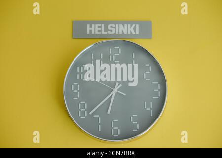 Un orologio elegante mostra l'ora contro una parete gialla vibrante, completata da un'elegante insegna che legge Helsinki sopra di essa. Foto Stock