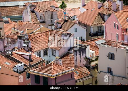 Tetti di edifici a Lisbona Portogallo // LISBONA, Portogallo — Un fitto gruppo di edifici residenziali con tetti tradizionali in terracotta riempie la cornice, mostrando il carattere architettonico di Lisbona. Gli edifici, dipinti in varie tonalità pastello, tra cui rosa e giallo pallido, presentano numerosi camini e antenne visibili, indicativi della vita urbana. Questa vista cattura il caratteristico paesaggio urbano della capitale portoghese, conosciuta per il suo terreno collinare e i quartieri storici. L'immagine fornisce una prospettiva ravvicinata dei tetti della città, evidenziando l'edificio comune ma Foto Stock
