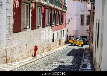 Tuk Tuk giallo su strada acciottolata a Lisbona Portogallo | LISBON — Un tuk-tuk giallo scende una ripida strada acciottolata nel centro storico di Lisbona, Portogallo, passando davanti a pedoni e un idrante rosso posizionato contro un muro di pietra. Questi veicoli a tre ruote, costruiti sulla piattaforma Piaggio Ape, sono diventati una vista comune per visite guidate attraverso i quartieri stretti e collinosi di Alfama e Bairro Alto a Lisbona. Foto Stock