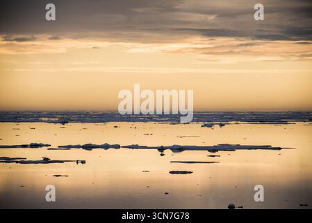 Klerckbukta Ice Floes Nordaustlandet Svalbard Norvegia // NORDEAUSTLANDET, Svalbard - i banchi di ghiaccio scivolano sulle acque calme di Klerckbukta sotto un cielo spettacolare. La scena cattura la dura bellezza del paesaggio artico, caratterizzato dalle sue vaste distese di ghiaccio e acqua. Nordaustlandet è la seconda isola più grande dell'arcipelago delle Svalbard, conosciuta per le sue ampie calotte di ghiaccio e la remota natura selvaggia. Questa regione è un habitat vitale per la fauna selvatica artica, anche se nessun animale è visibile in questa particolare immagine. Il basso angolo del sole, riflesso sulla superficie dell'acqua, crea un eth sereno e in qualche modo sereno Foto Stock