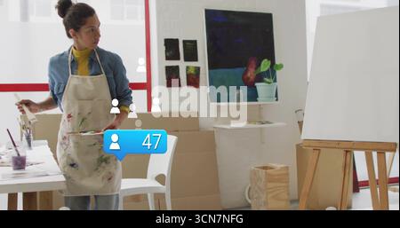 Artista matura che mescola la pittura su palette in uno studio creativo, con cavalletto e tele Foto Stock
