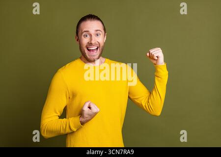 Giovane allegro con lunghi capelli biondi in camicia gialla celebra il successo su sfondo cachi esprimendo felicità Foto Stock