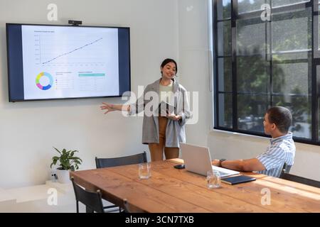 Colleghi afroamericani che puntano sullo schermo montato a parete mentre utilizzano tablet o laptop in ufficio Foto Stock