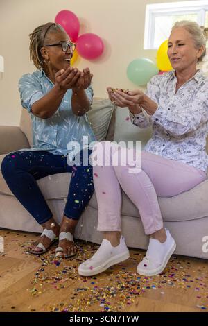 Diverse amiche anziane che lanciano coriandoli colorati sul divano di casa con palloncini color pastello Foto Stock
