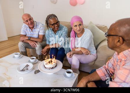 Diversi amici anziani festeggiano il compleanno a casa intorno al tavolino da caffè con torta e palloncini Foto Stock