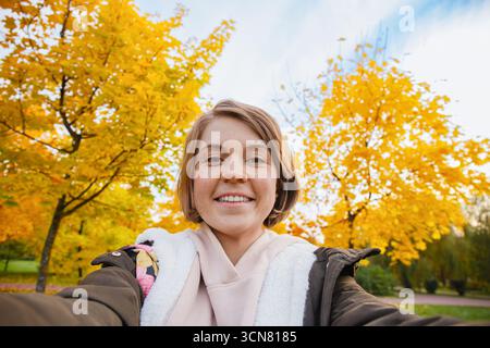 Donna sorridente con cappuccio e giacca tiene la fotocamera per i selfie, incorniciata da foglie d'acero dorato e cielo blu nel parco stagionale, stile di vita autunnale, all'aperto Foto Stock